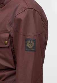Крупен план на ръкав на кафяво яке с лого на Belstaff и бутонен джоб на гърдите.