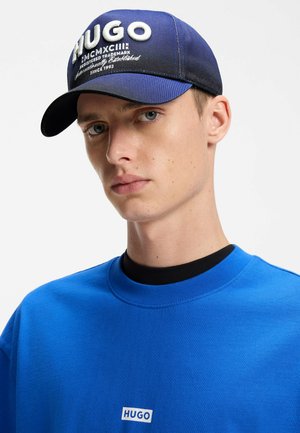 Junger Mann mit einer blauen HUGO-Mütze und passendem blauem HUGO-Sweatshirt vor einem schlichten weißen Hintergrund.