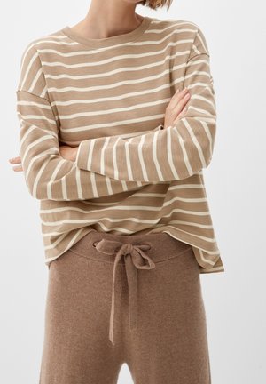 Osoba ubrana w beżową i białą bluzkę w paski z długimi rękawami, ze skrzyżowanymi ramionami, oraz w brązowe spodnie ze sznurkiem na tle jednolitej białej ściany.