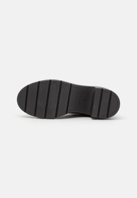 Suela de goma negra con surcos texturizados, que presenta un sutil logotipo de la marca. El diseño incluye crestas para tracción y estabilidad.