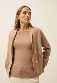 Suéter marrón de canalé con escote cuadrado, superpuesto a un cárdigan marrón claro con cuello de chal y cuatro botones.