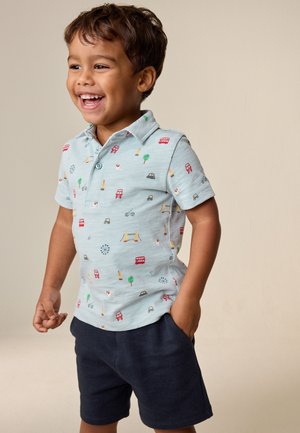 Lachende jonge jongen die een lichtblauw poloshirt met Londens thema opdruk en donkerblauwe korte broek draagt, staat met één hand in de zak.