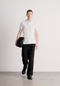 Λευκό πόλο πουκάμισο, μαύρο παντελόνι και μαύρα παπούτσια. Το μοντέλο κρατάει ένα σκούρο μπουφάν. Απλός, μοντέρνος σχεδιασμός με ουδέτερη παλέτα χρωμάτων.