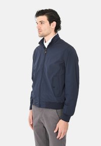 Giacca a zip di colore blu navy in tessuto leggero con colletto rialzato, vita elastica e dettagli con tasche laterali. Finitura opaca con texture liscia.