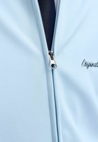 Ανοιχτό μπλε μπουφάν με φερμουάρ, μερικώς ανοιχτό που αποκαλύπτει σκοτεινό εσωτερικό στρώμα, με αχνό μαύρο κεντημένο κείμενο πάνω στο ύφασμα.