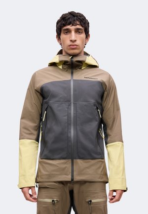 Man draagt een colour-blocked bruine, grijze en gele outdoor jas met capuchon, ritszakken en bijpassende bruine broek.