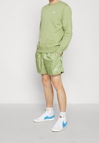 Zelená mikina se shodnými šortkami vyrobenými z lehkého materiálu, s bílým logem Nike. Doplněno bílými a modrými vysokými teniskami.