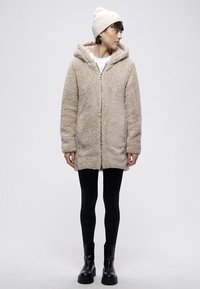 Bež plašč iz puhasto volnene tkanine s kapuco, sprednjo zadrgo in stranskimi žepi; kombiniran s črnimi legicami in robustnimi črnimi škornji.