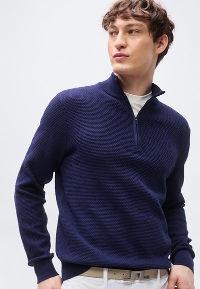 Νεαρός άνδρας που φοράει μπλε σκούρο πουλόβερ με φερμουάρ ως το στήθος πάνω από λευκό πουκάμισο, συνδυασμένο με λευκό παντελόνι και μπεζ ζώνη, κοιτάζοντας πλάγια.