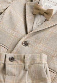 Costume beige clair avec un motif à carreaux dans des tons jaunes et orange. Comprend des boutons marron et un nœud papillon assorti. Texture de tissu douce.