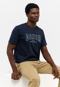 Tamsiai mėlyna medvilninė marškinėliai su "DENIM" grafiniu tekstu baltos ir šviesiai mėlynos spalvos. Apvalia iškirpte, trumpomis rankovėmis, derinami su bežiniais džinsais.