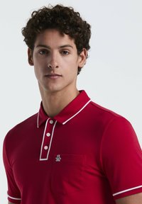 Polo rouge en coton, avec des accents blancs sur le col et la patte de boutonnage, comprenant une poche poitrine et un détail de logo.