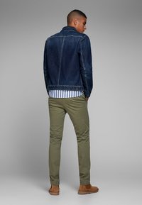 Denimjakke i mørkeblå, grønne bukser og en stribet skjorte. Brune ankelstøvler. Fokus på stoftekstur og pasform fra bagview.