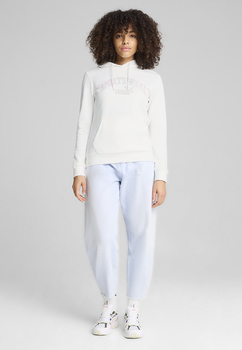 Joven con cabello rizado que lleva una sudadera deportiva blanca de Puma, jeans azul claro y zapatillas blancas, de pie contra un fondo liso.