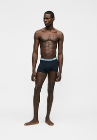 Atletski tamnoputi muškarac stoji bos nasuprot bijele pozadine, noseći tamnoplave bokserice Emporio Armani s svijetloplavim pojasom.