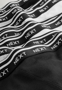 Černé a bílé bavlněné spodní prádlo s výrazným černým pasem a bílým textem "NEXT", zobrazené v vrstveném uspořádání.
