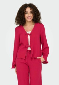 Blusa leggera e testurizzata di un brillante colore rosa, con scollo a V e dettagli con cordoncino sulla parte anteriore e sull'orlo, abbinata a pantaloni testurizzati coordinati.