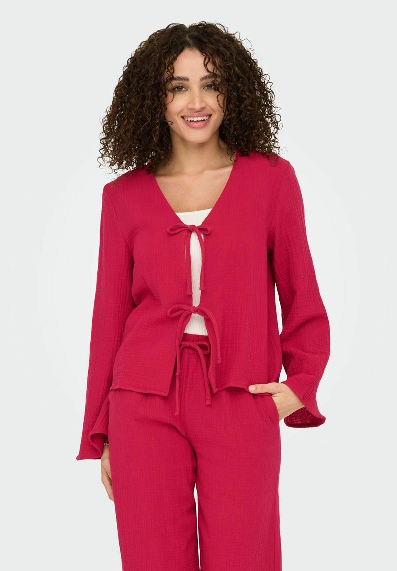 Blusa leggera e testurizzata di un brillante colore rosa, con scollo a V e dettagli con cordoncino sulla parte anteriore e sull'orlo, abbinata a pantaloni testurizzati coordinati.