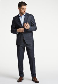 Abito a scacchi blu navy con giacca monopetto, camicia bianca e scarpe stringate marroni. Tessuto liscio, vestibilità su misura e design strutturato.