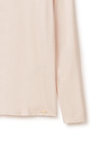 Světle růžová halenka s dlouhým rukávem vyrobená z jemné tkaniny, s prostřiženým lemem a malou zlatou značkovou nášivkou blízko spodního lemu.