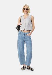 Chaleco corto gris claro con escote en V y botones plateados, combinado con jeans de tiro alto azul que presentan un patrón sutil y zapatos planos negros.