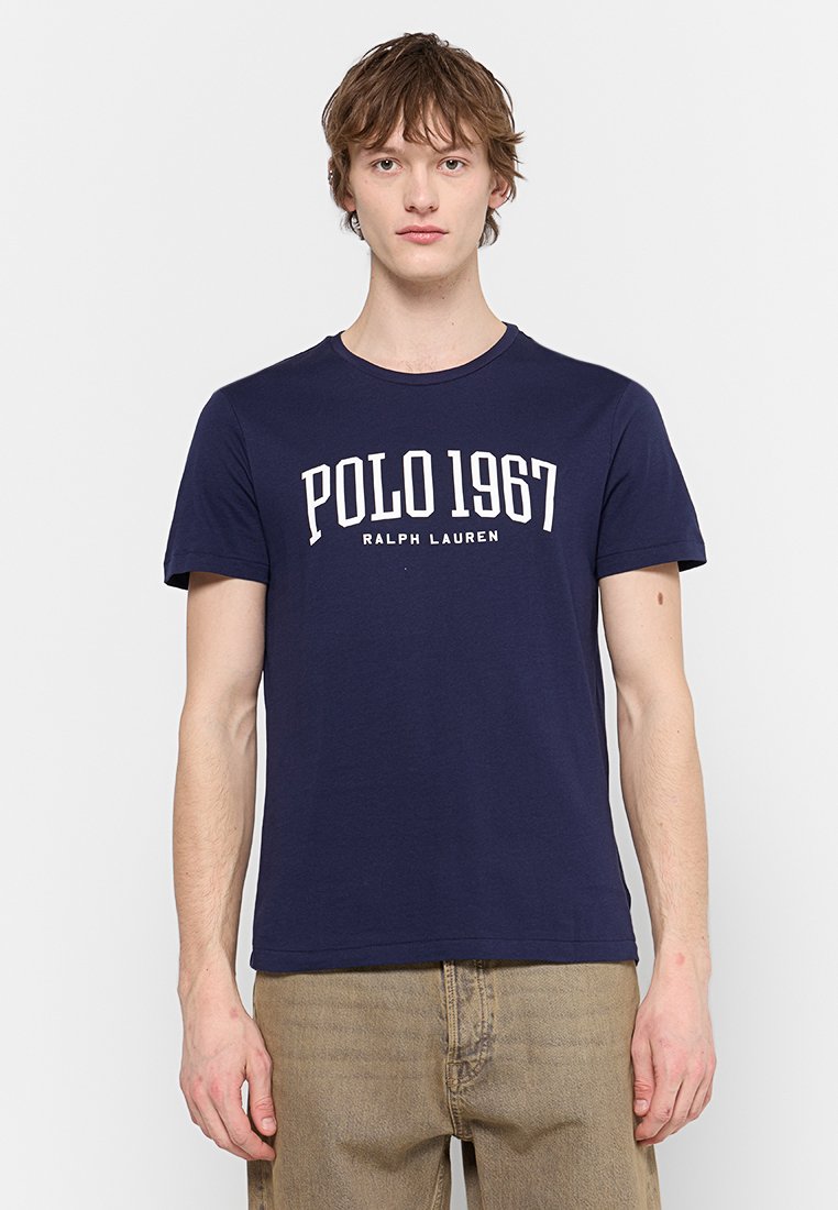 Giovane uomo che indossa una maglietta blu navy con la scritta bianca "POLO 1967 RALPH LAUREN" e pantaloni marrone chiaro, in piedi di fronte a uno sfondo neutro.