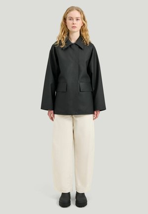 Kvinde med langt, krøllet hår iført en sort jakke, cremekarvede bukser med vide ben og sorte støvler mod en ensfarvet, lys baggrund.