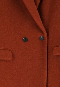 Manteau en laine couleur rouille avec une texture douce, doté de deux boutons noirs et d'un col cranté, ainsi que de deux poches à l'avant.