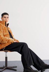 Chaqueta de piel sintética naranja con cremallera, camisa negra, vaqueros negros holgados y botas negras robustas. Persona sentada en una silla giratoria simple.