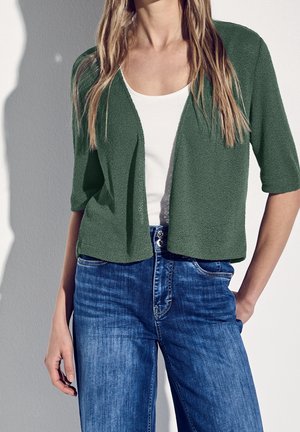 Kobieta w zielonym kardiganie na otwartym przodzie, na białym topie i niebieskich dżinsach z wysokim stanem, stoi z jedną ręką w kieszeni na jednolitym tle.