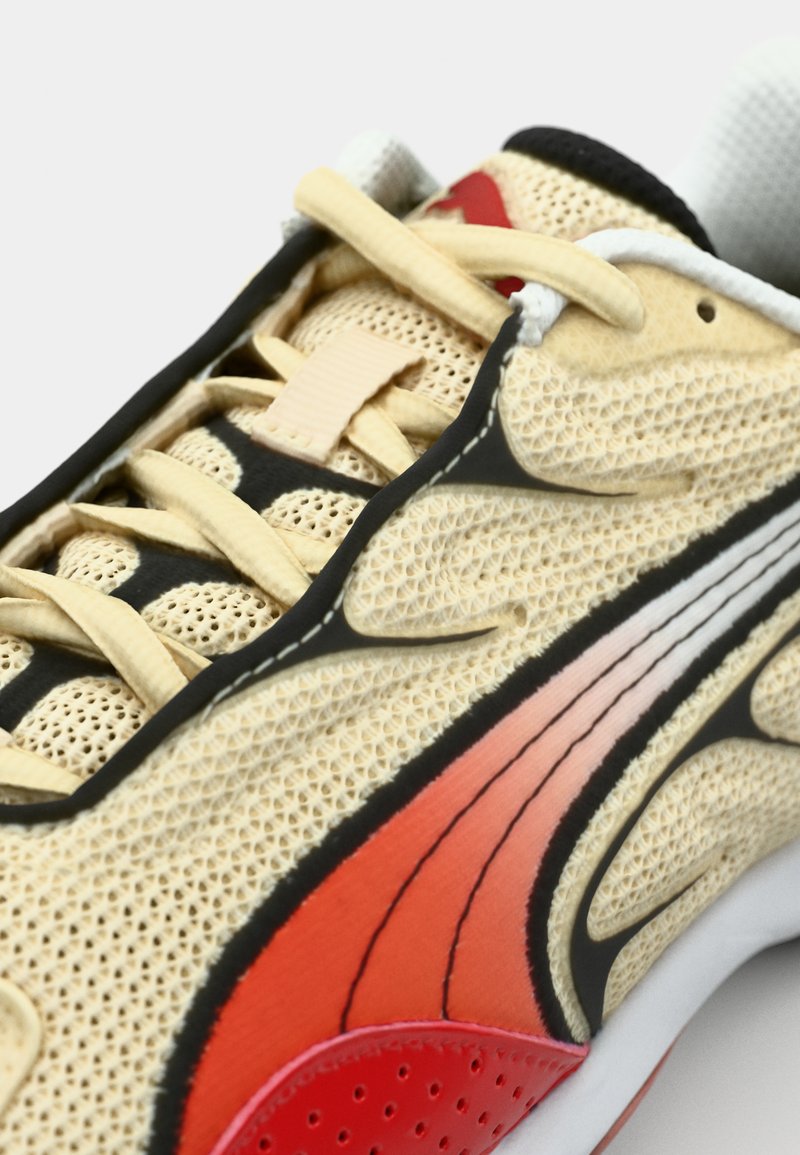 Chaussure de sport avec une tige en maille beige, accents noirs, détails rouges et oranges, et lacets beiges. Présente une surface texturée et des lignes de design courbées.