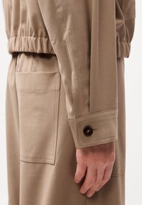 Gros plan du bras et de la main d'une personne portant un vêtement beige à manches longues avec poignet boutonné et grande poche sur le pantalon.
