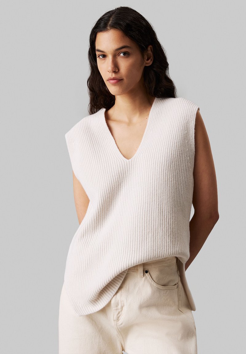 Femme avec de longs cheveux noirs portant un pull en tricot crème sans manches avec un décolleté en V et un pantalon taille haute beige clair, se tenant devant un fond gris uni.