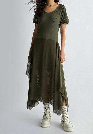 Kvinde iført olivengrøn kjole med korte ærmer og asymmetrisk gennemsigtigt skørt, kombineret med hvide snørebåndscombatstøvler.