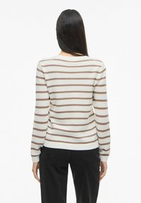 Femme aux cheveux longs et raides, portant un pull en maille rayé beige et marron et un pantalon noir, debout de dos sur fond blanc.