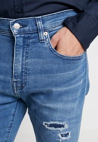Ljusblå denimjeans med hög midja, framfickor och slitna detaljer med fransade kanter. Knapp- och dragkedjestängning framtill.