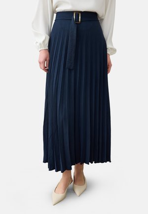Kvinde iført en navy plisseret midi nederdel med matchende bælte og beige spidse sko, stående mod en ensfarvet baggrund.