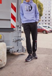 Felpa viola con logo e pantaloni della tuta neri con stampa del logo, abbinati a sneakers nere, in piedi vicino a un contenitore grigio.