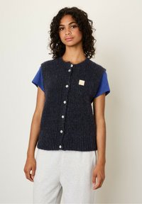 Marineblauwe textuur wolmix vest met een ronde hals, knoopsluiting aan de voorkant en een klein labelpatch, gedragen over een blauwe t-shirt en grijze broek.