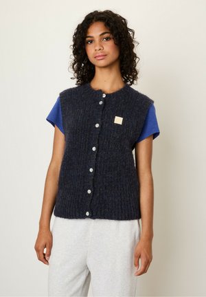 Gilet en mélange de laine texturée bleu marine avec un col rond, fermeture à boutons sur le devant et petite étiquette, porté sur un t-shirt bleu et un pantalon gris.