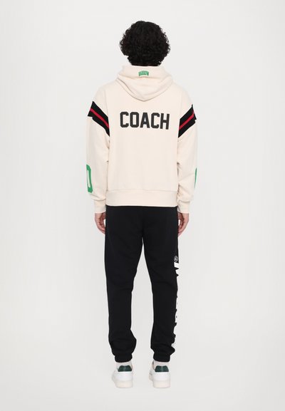 Hanorac crem cu "COACH" imprimat pe spate; are dungi roșii și negre pe mâneci. O pereche de pantaloni jogger negri cu imprimeu alb pe laterale.
