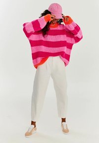 Růžový a oranžový pruhovaný pletený svetr, kombinovaný s bílými kalhotami a světlými růžovými teniskami. Svetr má oversized rukávy a žebrované manžety.