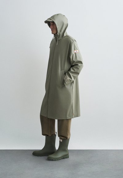 Homem de casaco de chuva verde com capuz, calças castanhas e botas de borracha verdes, de pé com as mãos nos bolsos do casaco contra um fundo simples.