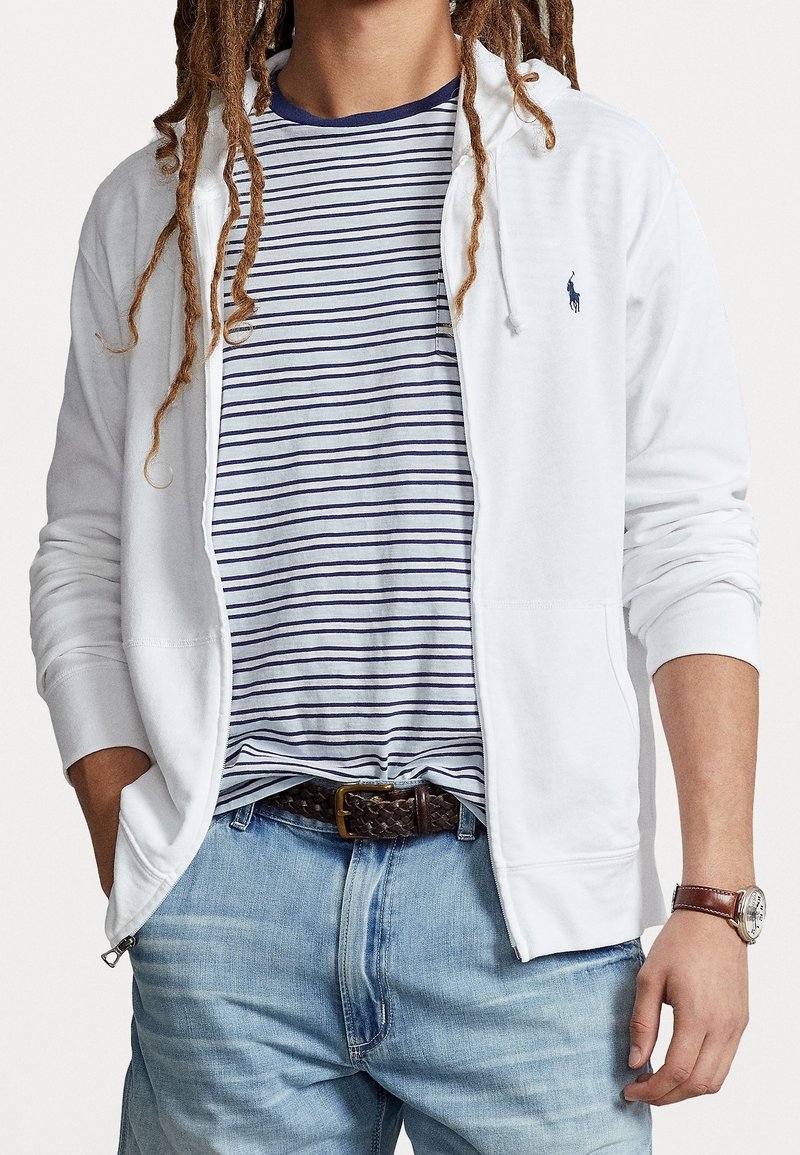 Weiße Zip-Up-Hoodie mit Kapuze, über einem navygestreiften T-Shirt getragen. Hellblaue Jeans und ein geflochtener brauner Gürtel vervollständigen das Outfit.