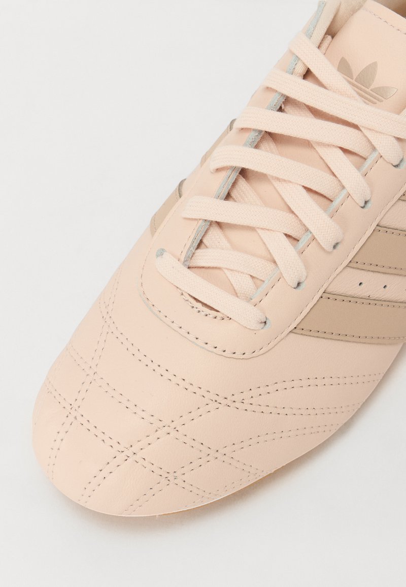 Zapato de cuero rosa claro con un patrón cosido, acentos beige y cordones claros. Presenta tres rayas paralelas en el lateral.