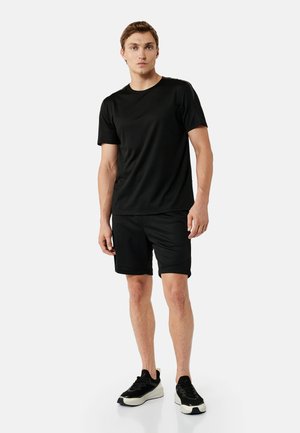 Mężczyzna stojący, ubrany w czarną koszulkę z krótkim rękawem, czarne szorty i czarne sneakersy z białymi podeszwami na jednolitym jasnym tle.