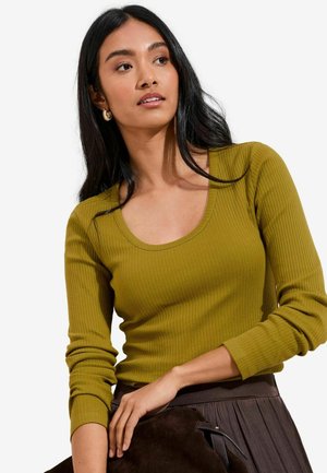 Femme aux longs cheveux noirs portant des boucles d'oreilles créoles en or, un haut à manches longues côtelé vert olive, tenant un sac en daim marron foncé.