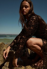 Abito in pizzo nero con maniche lunghe e svasate e motivo floreale. Modella accovacciata su rocce, oceano sullo sfondo, sotto un cielo blu chiaro.