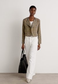 In het beige en bruin gekleurde croptop met luipaardprint, gecombineerd met een wit topje en wijdvallende witte broek. Het model houdt een zwarte tote bag vast en draagt lichte hakken.