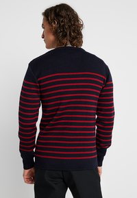 Granatowy sweter z poziomymi czerwonymi paskami, ściągacze na rękawach i na dekolcie, średnia faktura dzianiny, klasyczny krój i jednolita tylna część.
